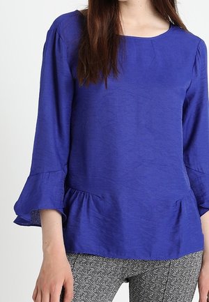 Blaue Bluse mit langen Ärmeln und quadratischem Ausschnitt, gerüschtem Taillensaum und strukturiertem Stoff. Kombiniert mit gemusterten Hosen.
