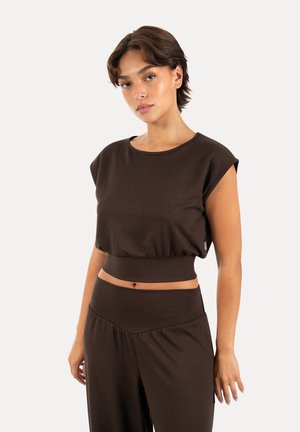 Ung kvinde med kort hår iført en mørkebrun ærmeløs crop top og matchende højttaljede bukser, der står mod en ensfarvet baggrund.