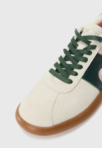 Beige suede sneaker with green laces and a dark green leather accent, featuring a pink detail and a brown rubber sole.