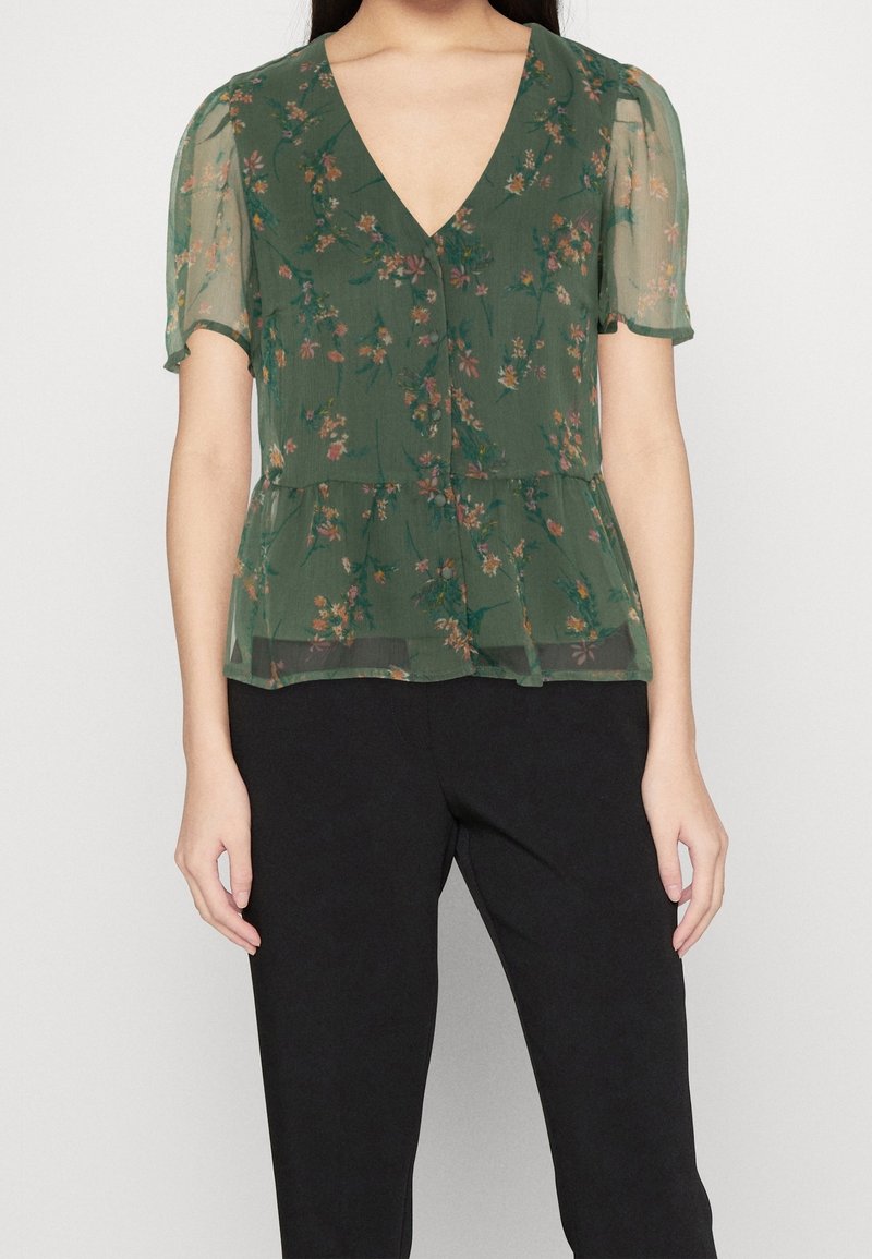 Haut transparent vert foncé à motif floral, manches courtes et encolure en V. Le design péplum ajoute un contraste structuré au tissu fluide.