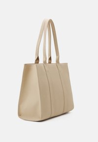 Beige-kangaskassi, jossa on rakenteellinen muotoilu, valmistettu teksturoidusta materiaalista. Kassissa on kaksi pitkää kantokahvaa ja pysty saumat lisäyksinä.