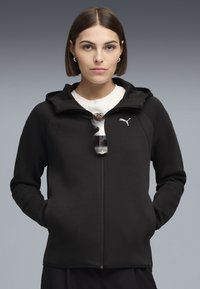 Giovane donna con capelli castani corti che indossa una felpa nera Puma con zip sopra una maglietta bianca, occhiali da sole appesi alla cerniera, mani nelle tasche.