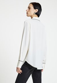 White long-sleeve shirt with a soft texture, black piping accents on cuffs, and a curved hem, paired with dark jeans.