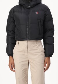 Giacca puffer nera cropped con zip frontale e logo Tommy Jeans abbinata a pantaloni beige a vita alta indossati da una persona.