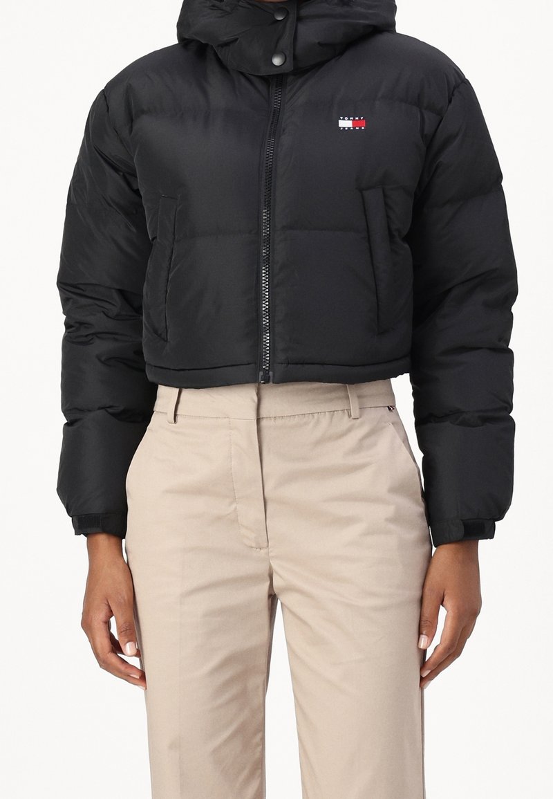 Giacca puffer nera cropped con zip frontale e logo Tommy Jeans abbinata a pantaloni beige a vita alta indossati da una persona.