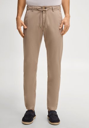 Beżowe spodnie o dopasowanym kroju z troczkiem i zapięciem na guzik, noszone z czarnymi wsuwanymi mokasynami na modelce stojącej.