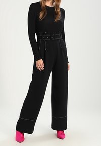 Pantalones de pierna ancha negros con un dobladillo cosido en contraste y una cintura con cinturón. Combinados con una blusa negra ajustada y tacones rosas.