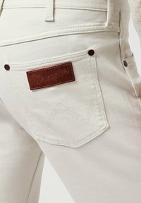 Jeans in denim beige chiaro con un patch in pelle marrone Wrangler, due tasche posteriori con cuciture decorative e accessori in metallo.
