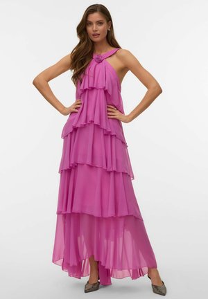 Kvinne iført en ermeløs, lagdelt rosa chiffonkjole med blomsterdetalj ved halsen, poserer med hendene på hoftene og sølvspisse sko.