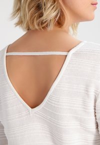 White knit top featuring textured stripes, a deep V-back design, and a narrow strap across the upper back. Soft, lightweight material.