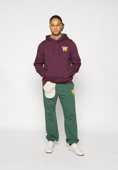 Vinröd huvtröja med gult logotyp, parat med gröna sweatpants med matchande logotyp, krämfärgad keps på byxorna och vita sneakers.