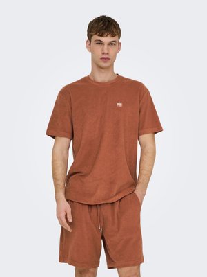 Jeune homme portant une chemise à manches courtes et un short en tissu éponge couleur rouille assortis, debout devant un fond clair uni.