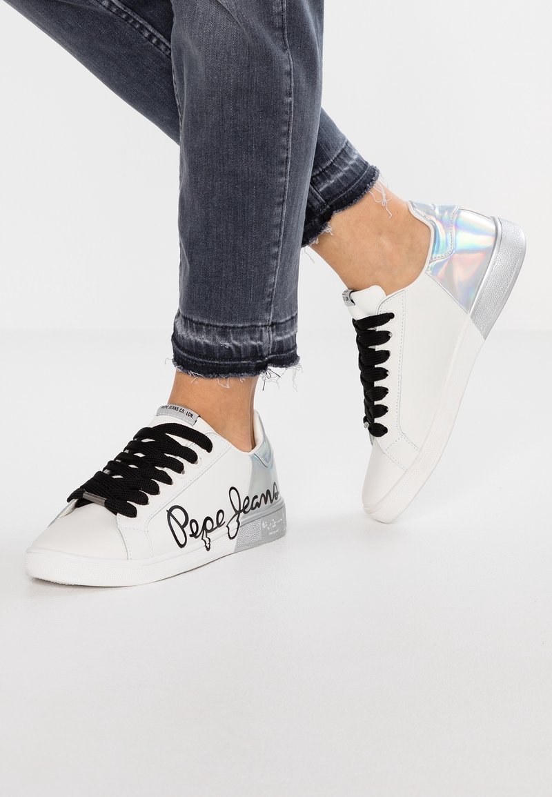 Baskets blanches avec lacets noirs et détail holographique au talon, portées avec un jean en denim gris foncé effiloché.