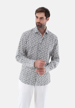Weißes Hemd mit Knopfleiste, das ein schwarzes Blumenmuster, kontrastierende Manschetten und einen klassischen Kragen aufweist. Getragen mit heller Hose.