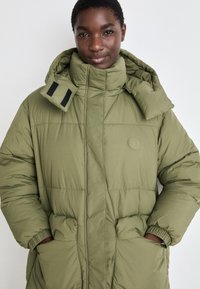 Olive green puffer coat with a high collar, large pockets, and elastic cuffs; features a logo and adjustable hood with Velcro accents.