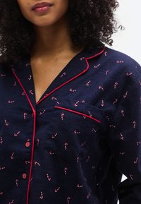 Haut de pyjama en flanelle marine avec motif de cannes en sucre d'orge rouges, passepoil rouge contrastant, poche poitrine et fermeture à boutons. Texture douce.