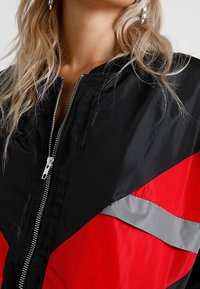 Giacca a vento nera e rossa caratterizzata da una chiusura con zip, dettagli in argento e un design geometrico con pannelli a contrasto. Texture liscia.