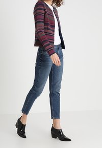 Strickjacke mit einem geometrischen Muster in Dunkelblau, Pink und Creme, kombiniert mit eng anliegenden blauen Jeans und schwarzen Lackstiefeln mit Absatz.