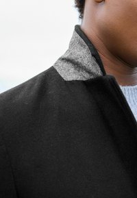 Abrigo de lana negro con un cuello de fieltro gris. La textura es suave con un diseño estructurado, costuras visibles y un cuello erguido.