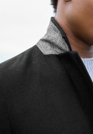 Abrigo de lana negro con un cuello de fieltro gris. La textura es suave con un diseño estructurado, costuras visibles y un cuello erguido.