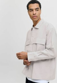 Uomo con tatuaggi che indossa una camicia a quadri grigia chiara, guardando di lato su uno sfondo bianco uniforme.