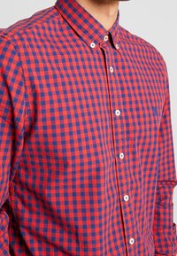 Camicia a quadri rossa e blu con colletto, bottoni bianchi e un piccolo logo sul petto, dal tessuto morbido.