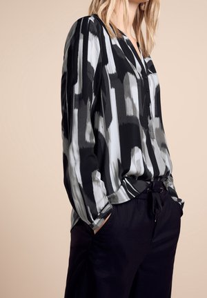 Vrouw draagt een blouse met lange mouwen met zwart-wit-grijze abstracte strepen en knopen, gecombineerd met een zwarte broek, handen in de zakken, beige achtergrond.