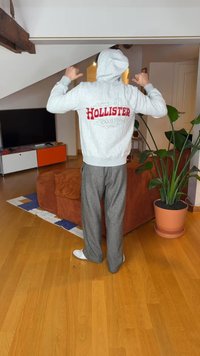 Szürke kapucnis pulóver piros „HOLLISTER” felirattal és logóval, szürke kockás nadrággal párosítva, fa padlón állva, zöld növény és kanapé mellett.