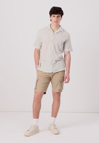 Camisa de botones de manga corta de color claro con un patrón ondulado, combinada con pantalones cortos de cargo color beige que tienen bolsillos laterales y un diseño sencillo.