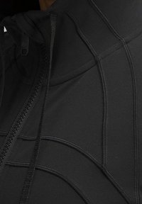 Detailní záběr na černou mikinu s kapucí, která má zip, nastavitelné šňůrky a precizní prošívání na látce.