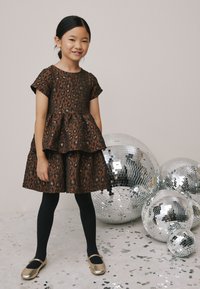 Barna leopárdmintás ruha réteges szoknyával, rövid ujjakkal és texturált felülettel. Fekete harisnyával és arany, kerek orrú cipővel párosítva.