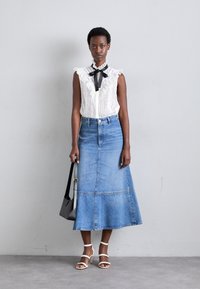 Hvid ærmeløs bluse med flæset krave, kombineret med en blå denim A-formet nederdel. Tilbehør inkluderer en sort håndtaske og hvide hælede sandaler.