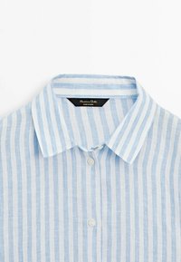 Camicia di lino a righe azzurro chiaro e bianco con colletto appuntito, caratterizzata da una patta frontale con bottoni e un'etichetta del marchio all'interno del colletto.