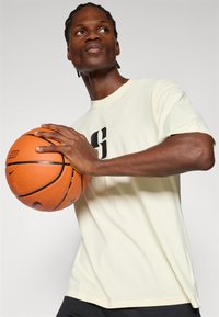 Helelilla puuvillane T-särk musta graafika kujundusega, hoides samal ajal oranži korvpalli. Taust on neutraalne.