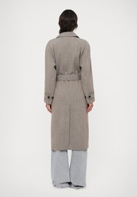 Beige kostkovaný trenčkot s páskem v pase, dlouhými rukávy a velkými knoflíky. Nositý přes šedé široké kalhoty.