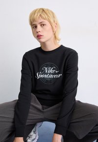 Črna mikica s srebrnim logotipom "Nike Sportswear" na prsih. Model nosi sivo široke hlače. Gladek material; podrobnost s krožnim dizajnom.