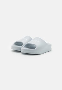 Chanclas de goma de color azul claro con punta abierta, que cuentan con una amplia tira y un interior texturizado. Logo de Lacoste en blanco en la superficie superior.