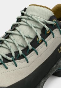 Zapato de senderismo gris claro con material texturizado, cordones elásticos en verde y detalles en dorado, y una suela de goma negra con un diseño estampado.