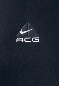 Black fabric features an embroidered triangular Nike logo and "ACG" in white text, showcasing a smooth texture and minimalist design.