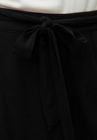 Ceinture en tissu noir avec un détail de nœud noué. La texture est légèrement côtelée, ajoutant un intérêt visuel au design.