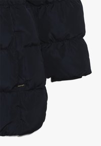 Manga y lado de una chaqueta acolchada negra que muestran costuras acolchadas y un bolsillo lateral parcialmente visible con una etiqueta de metal.