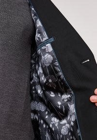 Blazer nero con fodera grigia texturizzata. L'interno presenta un motivo floreale in tonalità di grigio e nero, con un bordo blu a contrasto.