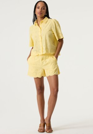 Vrouw met gevlochten haar, gekleed in een bijpassend geel ooglet-shirt met korte mouwen en een short met geschulpte randen en beige sandalen met hakken.