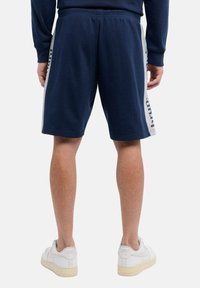 Man in marineblauwe shorts met lichtgrijze zijkanten en witte Nike Air sneakers, gezien vanaf de achterkant tegen een effen achtergrond.