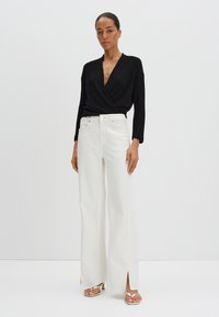 Zwarte geribbelde langemouwen wikkel top met een diepe V-hals en witte wijde broek met zijsplitsingen. Model draagt zilveren strappy hakken.