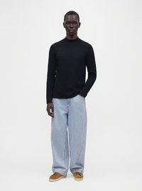 Homme portant une chemise noire à manches longues, un jean large bleu clair et des chaussures marron, debout devant un fond blanc uni.