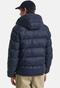 Mørkebl�å pufferjakke med hætte, der har et quiltet design og tyk isolering. Blød, glat tekstur med elastiske manchetter og kant.