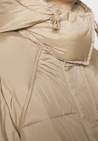 Beige pufferjakke med høj krave, justerbare snore og en glat, blank overflade med synlig quiltet syning.