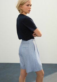Pantaloni corti in chambray blu con una parte anteriore plissettata e dettagli sulla fascia in vita, abbinati a una maglietta scura a maniche corte, osservati di lato.