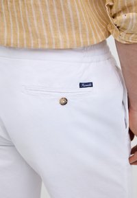 Pantalon blanc en coton avec une poche arrière dotée d'un bouton et d'une petite étiquette bleue. Le tissu a une texture lisse.